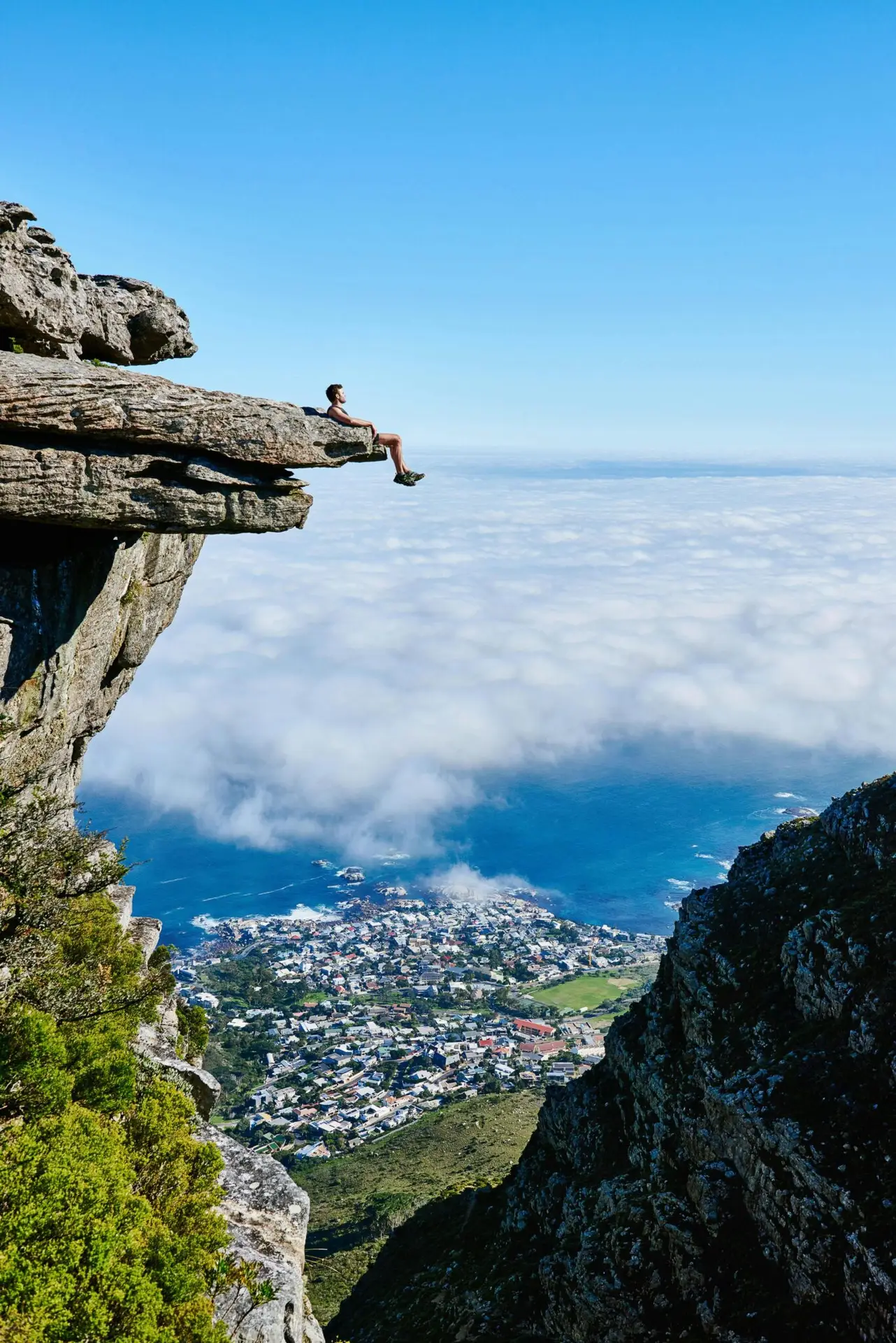 A person sits on a cliff edge enjoying a breathtaking view of the ocean and city below.